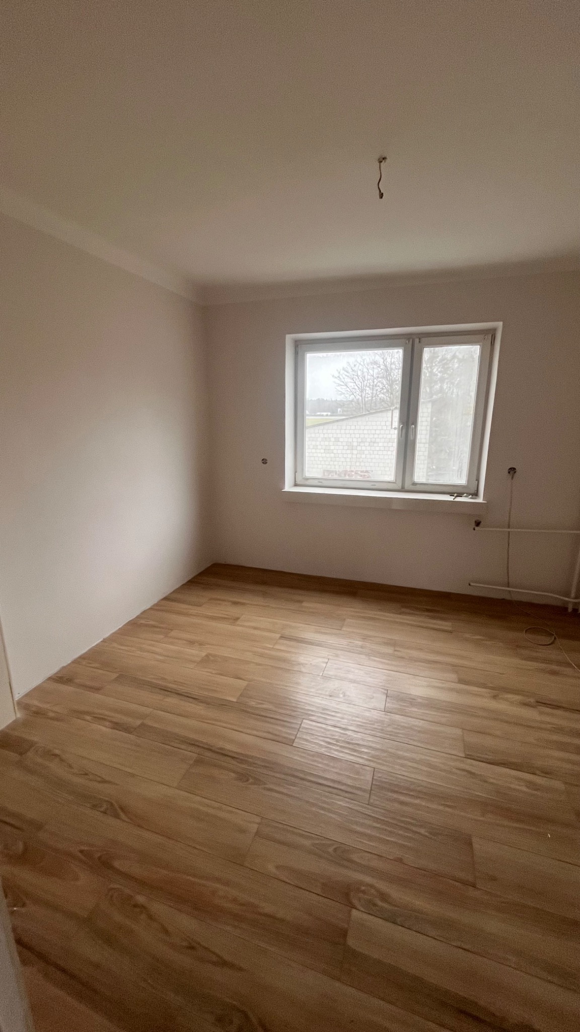 Pusty pokój po remoncie z nową drewnopodobną podłogą, świeżo pomalowanymi ścianami i oknem z widokiem na budynek gospodarczy.