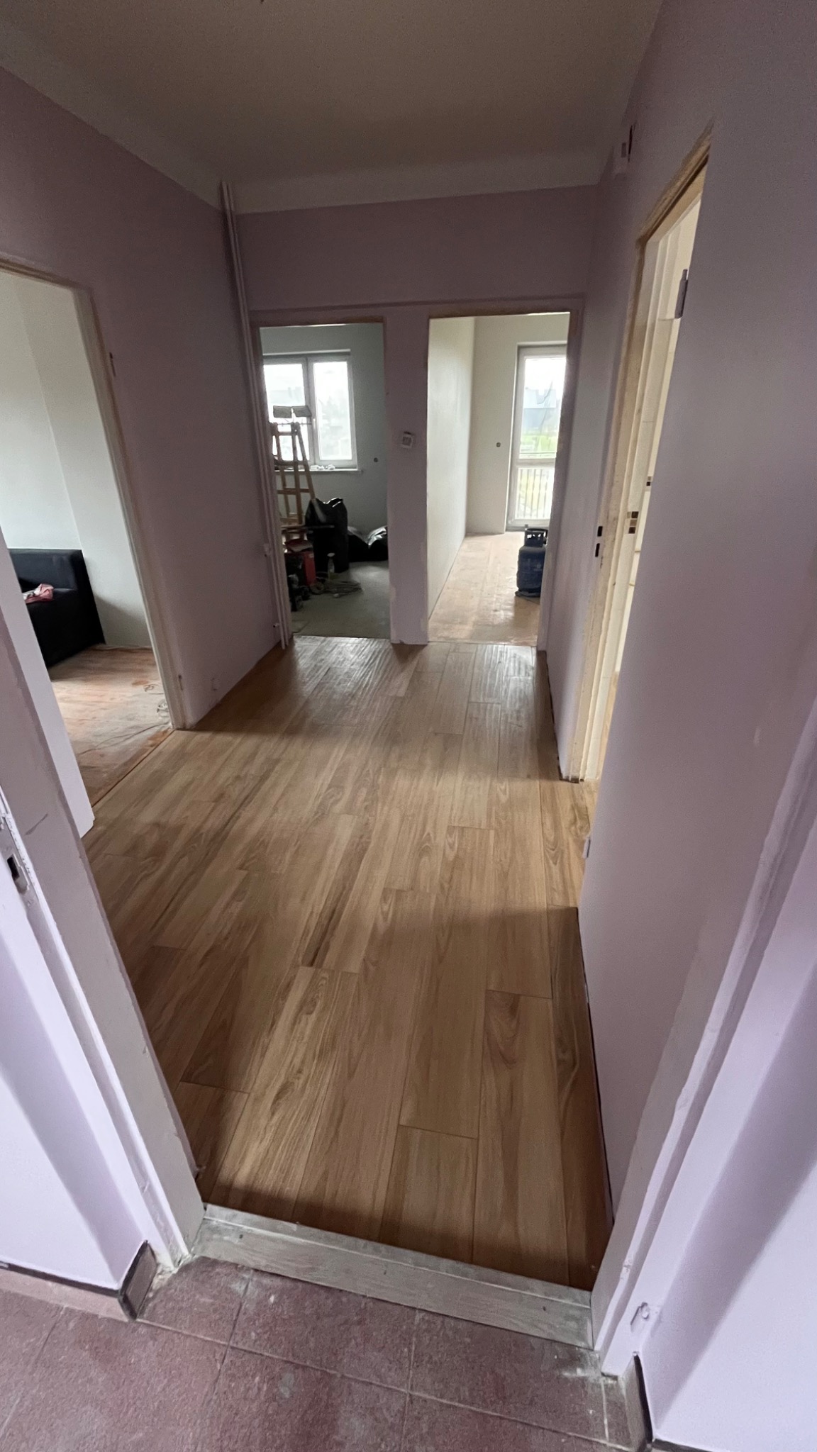 Korytarz w trakcie remontu, widoczne nowe panele podłogowe imitujące drewno, ściany pomalowane na jasny fiolet, w tle widoczne kolejne pomieszczenia z elementami rusztowania i narzędzi.