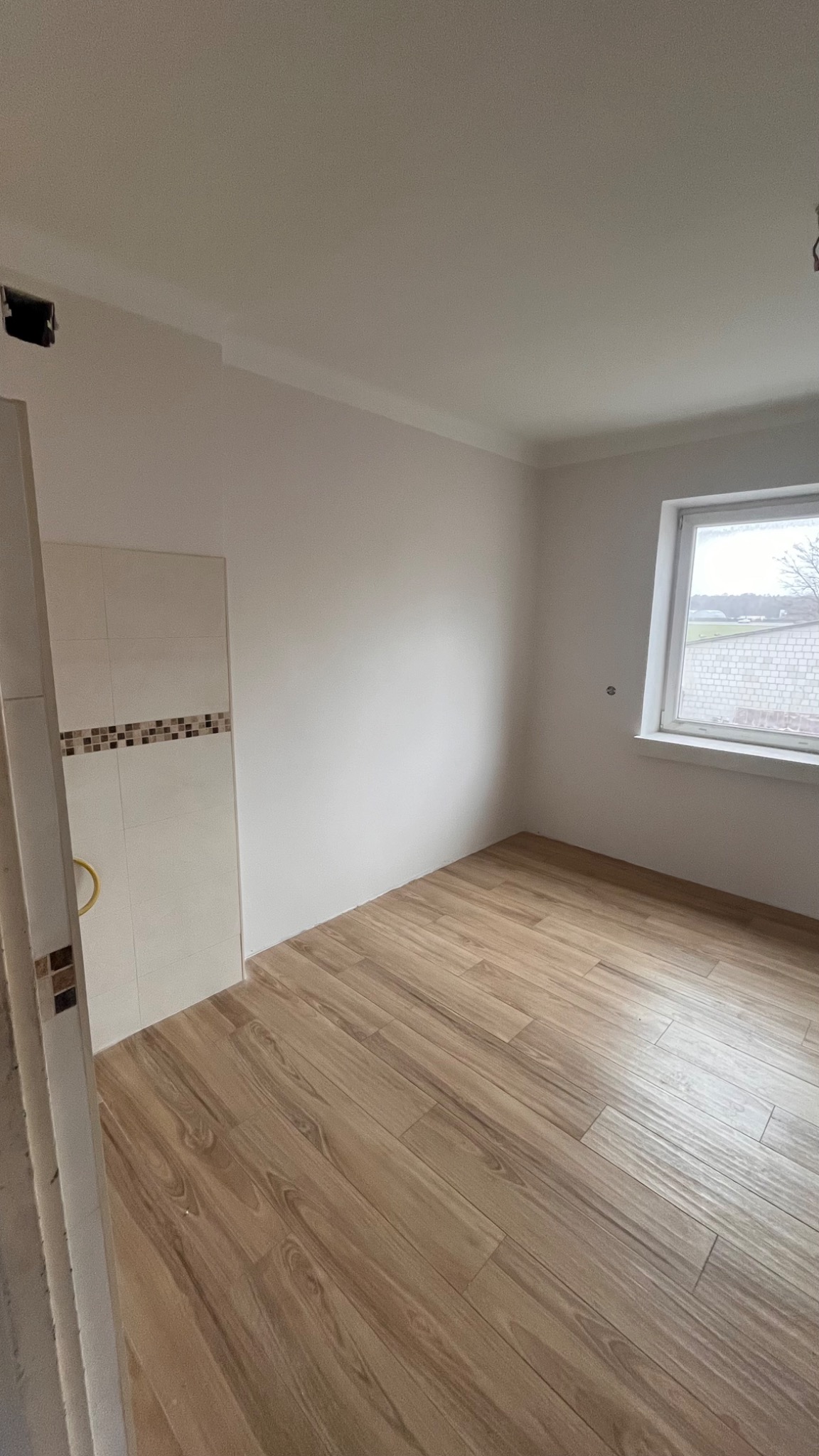 Pusty pokój po remoncie z panelami podłogowymi imitującymi drewno, białe ściany i okno z widokiem na budynek gospodarczy.