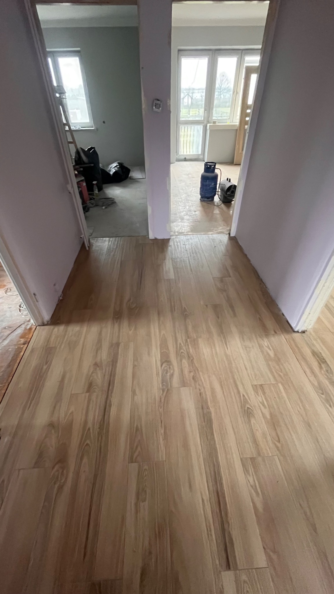 Ułożona podłoga z paneli imitujących drewno, widok z korytarza na dwa pomieszczenia w trakcie remontu, w jednym drabina i worki, w drugim butla gazowa i nagrzewnica.