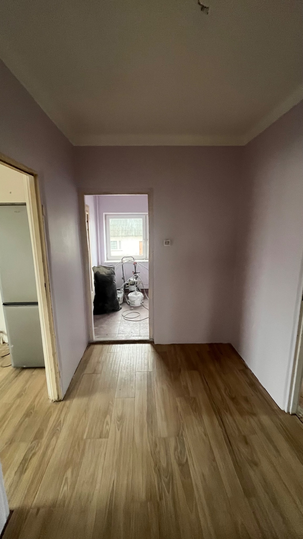 Wnętrze mieszkania w trakcie malowania: ściany pomalowane na jasny fiolet, widoczny sprzęt do malowania natryskowego i worki z materiałami w pokoju obok, jasne panele podłogowe.