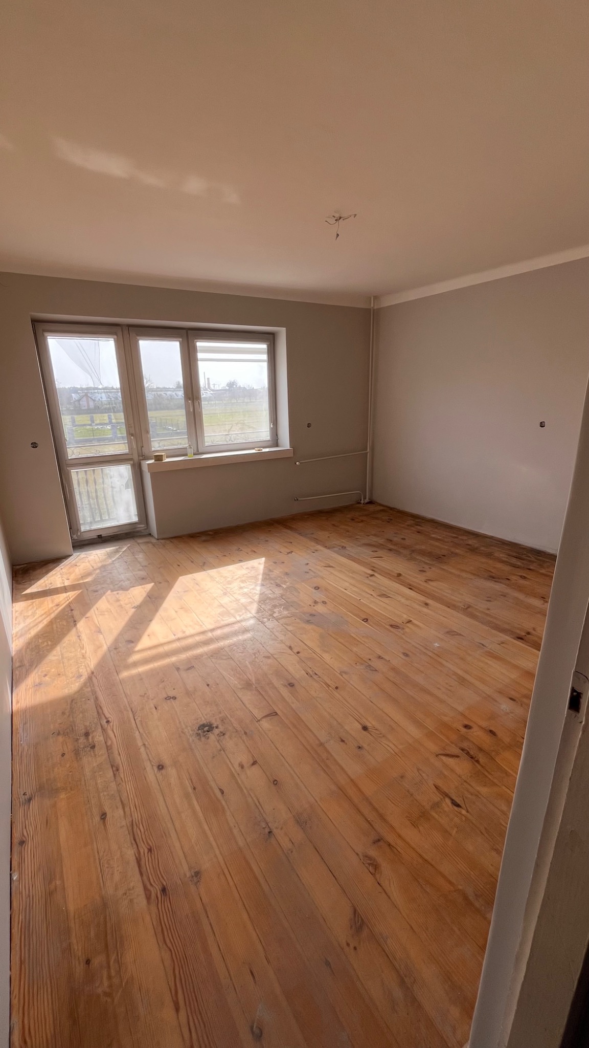Pusty pokój z drewnianą podłogą, szarymi ścianami i jasnym oknem, widoczne wystające kable sufitowe. Podłoga z widocznymi śladami użytkowania.