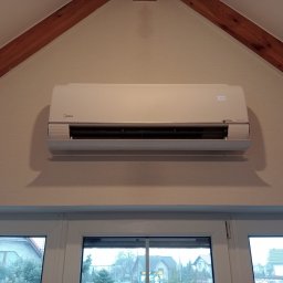 PRZEDSIĘBIORSTWO HANDLOWO-USŁUGOWE SZOPIŃSCY SPÓŁKA Z OGRANICZONĄ ODPOWIEDZIALNOŚCIĄ - Biały klimatyzator Midea zamontowany w domu na poddaszu, widoczny na tle drewnianych belek stropowych i okna z widokiem na zabudowę jednorodzinną.