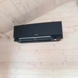 Czarna jednostka wewnętrzna klimatyzatora Daikin zamontowana na jasnej, drewnianej ścianie w pomieszczeniu o skośnym suficie wykończonym boazerią.