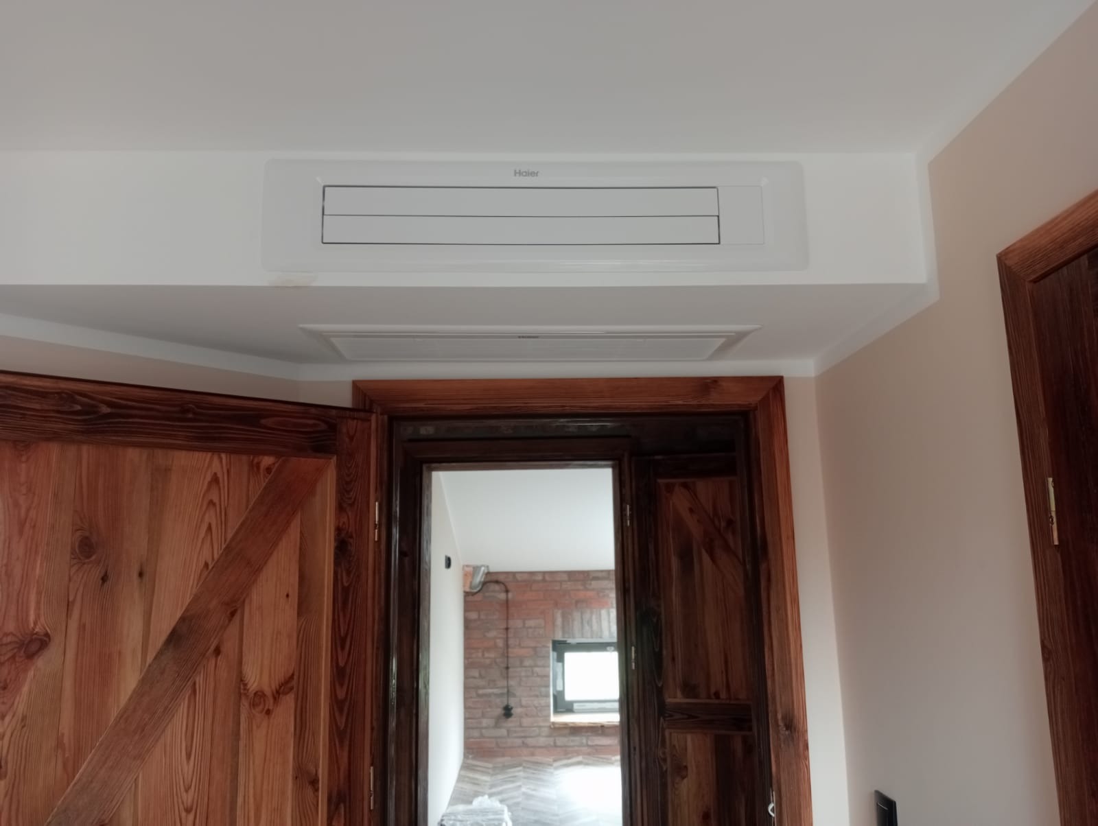 Biała, podwieszana jednostka klimatyzacyjna Haier zamontowana na suficie w pomieszczeniu z drewnianymi drzwiami w stylu rustykalnym i widoczną ścianą z czerwonej cegły w tle.
