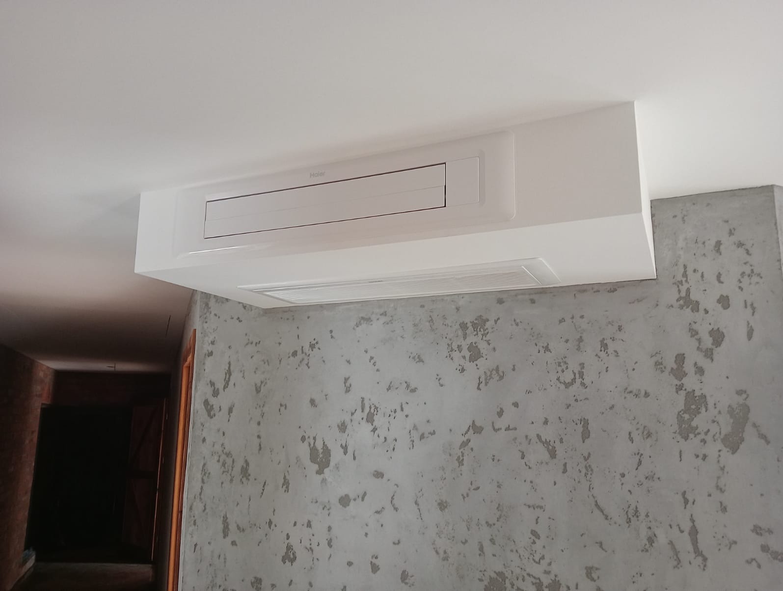 Biała klimatyzacja kasetonowa Haier zamontowana w podwieszanym suficie, widoczna szara ściana z efektem betonu architektonicznego.