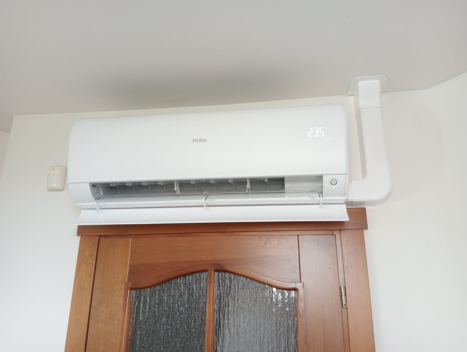 Biały klimatyzator Haier zamontowany nad drewnianymi drzwiami z szybą, widoczny wyświetlacz z temperaturą 23.5 stopnia Celsjusza, biała rura odprowadzająca skropliny.