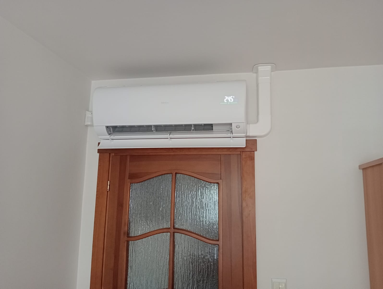 Biały klimatyzator zamontowany nad drewnianymi drzwiami z szybami, wyświetlający temperaturę 24.5 stopnia Celsjusza, z widocznymi przewodami odprowadzającymi.