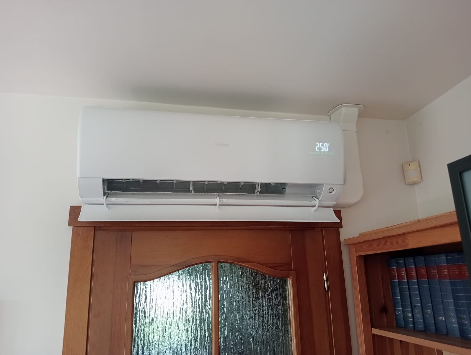 Biały klimatyzator Haier zamontowany nad drewnianymi drzwiami z szybą, wyświetlający temperaturę 25 stopni Celsjusza, obok regał z książkami.