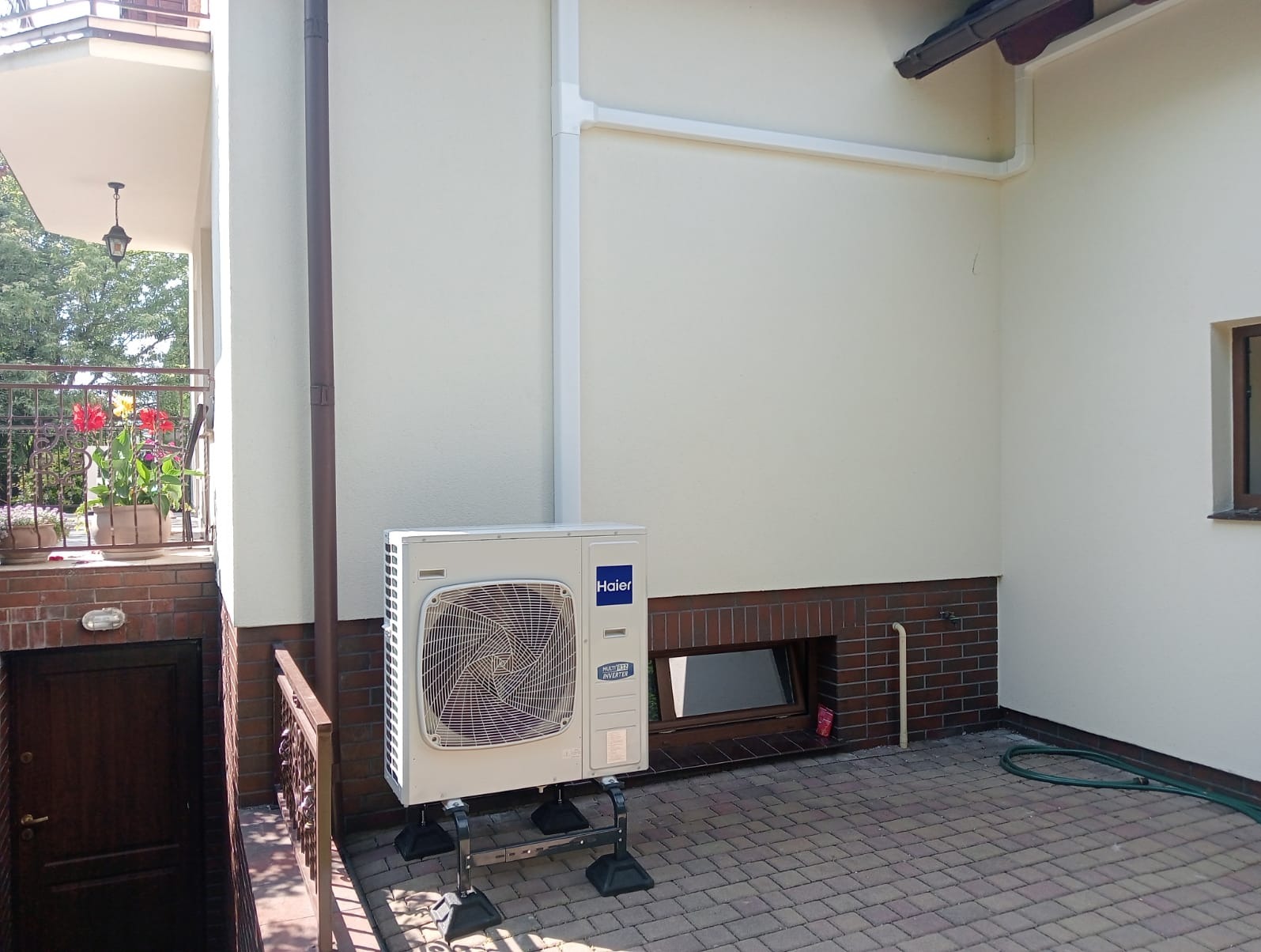 Zewnętrzna jednostka klimatyzacji Haier zamontowana na specjalnej konstrukcji na kostce brukowej, widok z boku domu z kwiatami na balkonie.