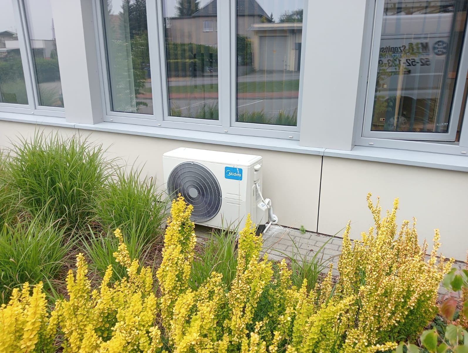 Biała jednostka zewnętrzna klimatyzacji Midea zamontowana przy ścianie budynku z szarymi oknami, otoczona dekoracyjną roślinnością z przewagą żółtych krzewów.