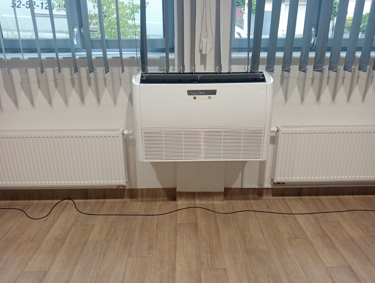 Biała klimatyzacja przypodłogowa Midea zamontowana w biurze, widoczne grzejniki i żaluzje okienne.