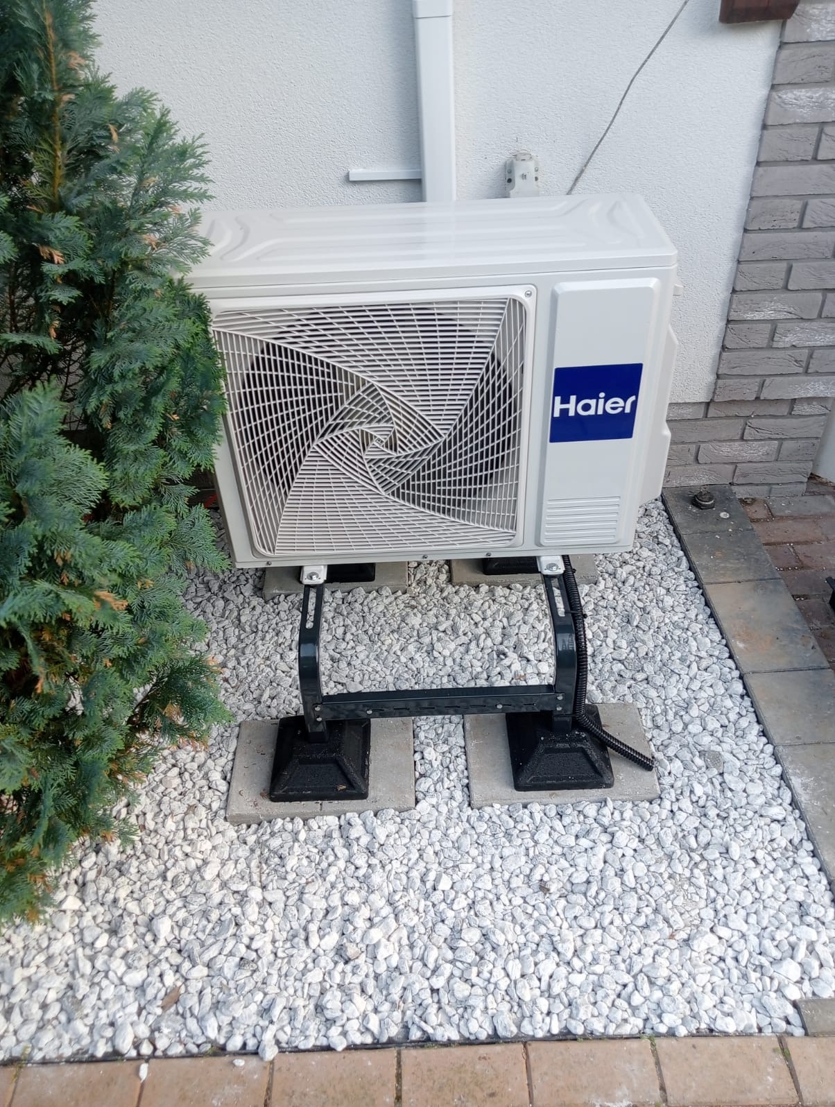 Zewnętrzna jednostka klimatyzacji Haier zamontowana na wspornikach na tle białego żwiru i ściany budynku, obok iglastego krzewu.