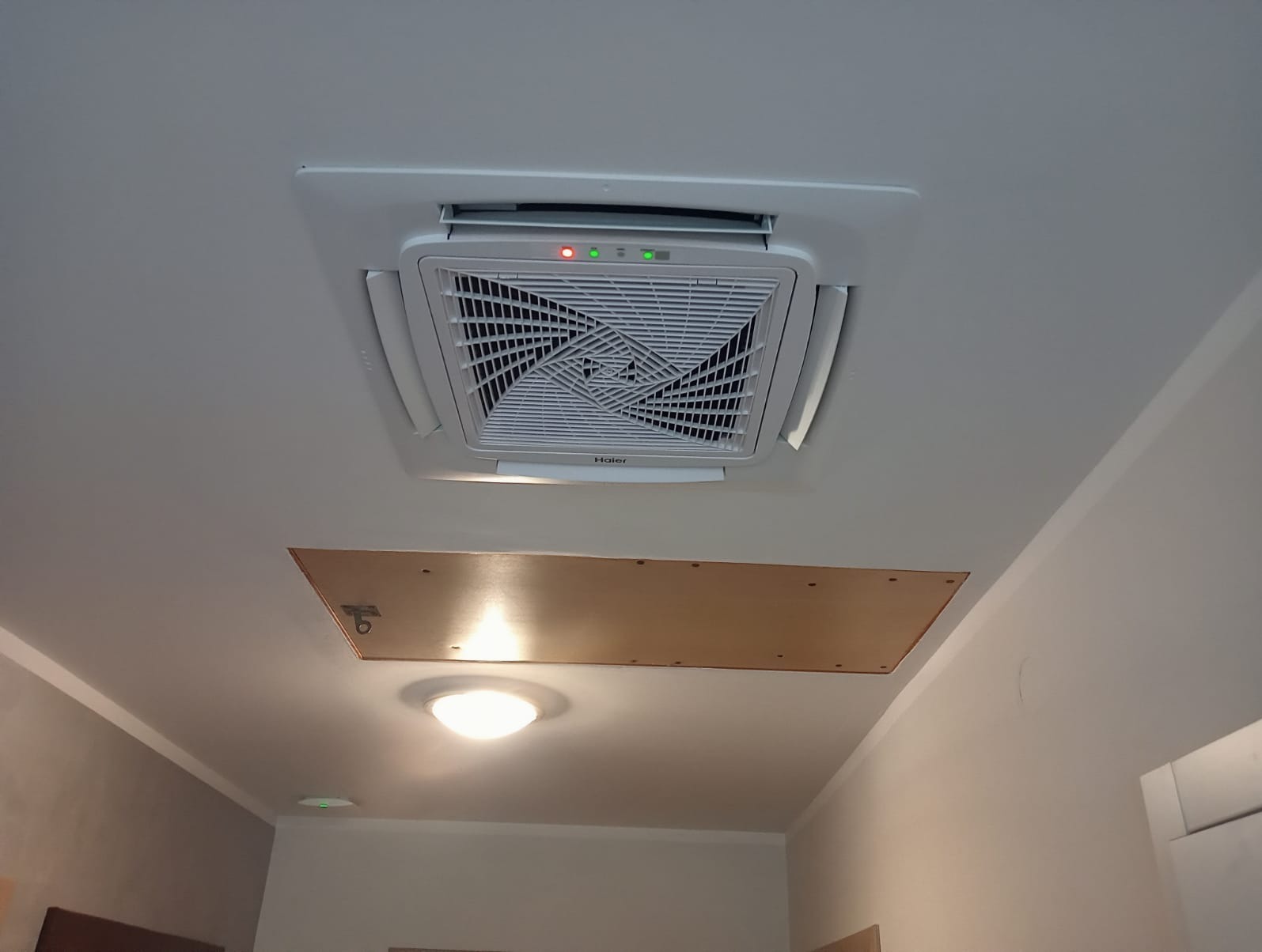 Sufitowy klimatyzator Haier z widocznymi diodami LED, zamontowany w pomieszczeniu z otworem rewizyjnym w suficie i lampą