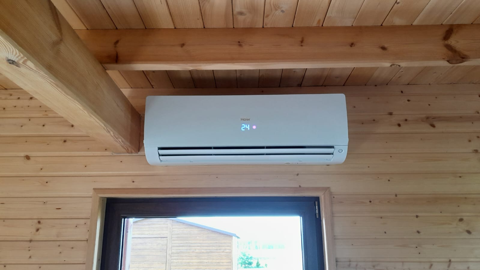 Biała klimatyzacja Haier zamontowana w drewnianym domu, wyświetlająca temperaturę 24 stopnie Celsjusza.