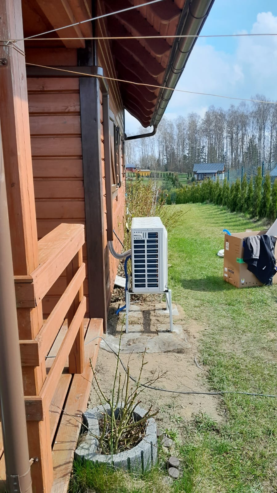 Zewnętrzna jednostka klimatyzacji Haier na metalowym stelażu, zainstalowana przy drewnianym domu z brązową stolarką, widoczny kabel i rurka łącząca z jednostką wewnętrzną, w tle trawnik i drzewa.