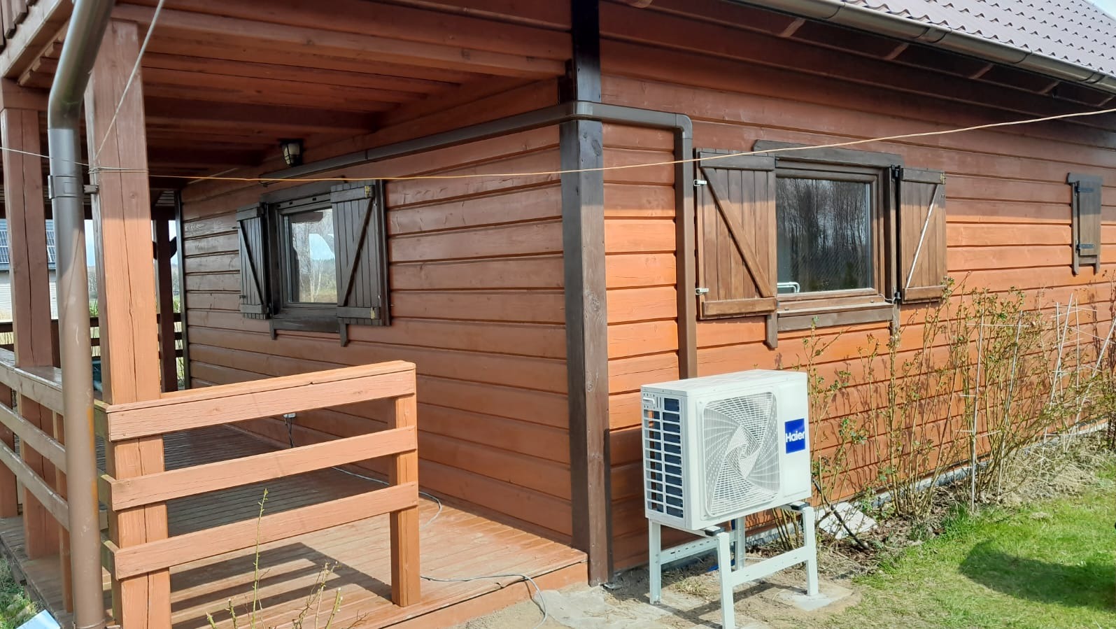 Zewnętrzna jednostka klimatyzacji marki Haier zamontowana na metalowym stelażu przy drewnianym, brązowym domu z okiennicami.