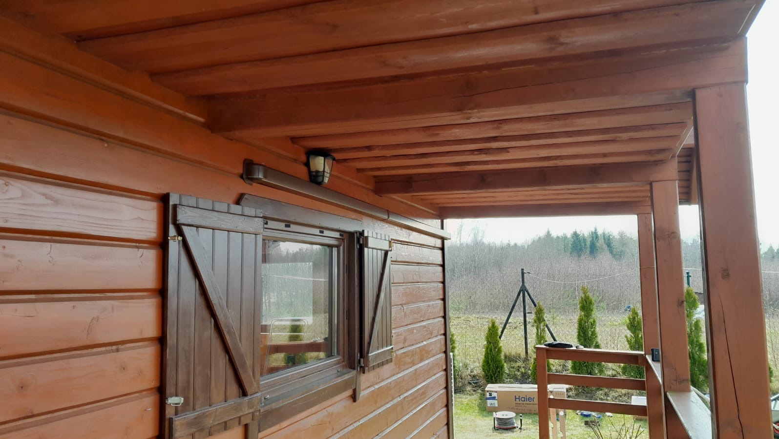 Drewniana elewacja domu z oknem i okiennicami, widoczny zewnętrzny moduł klimatyzacji i lampa na ścianie, zadaszony taras z widokiem na ogród.