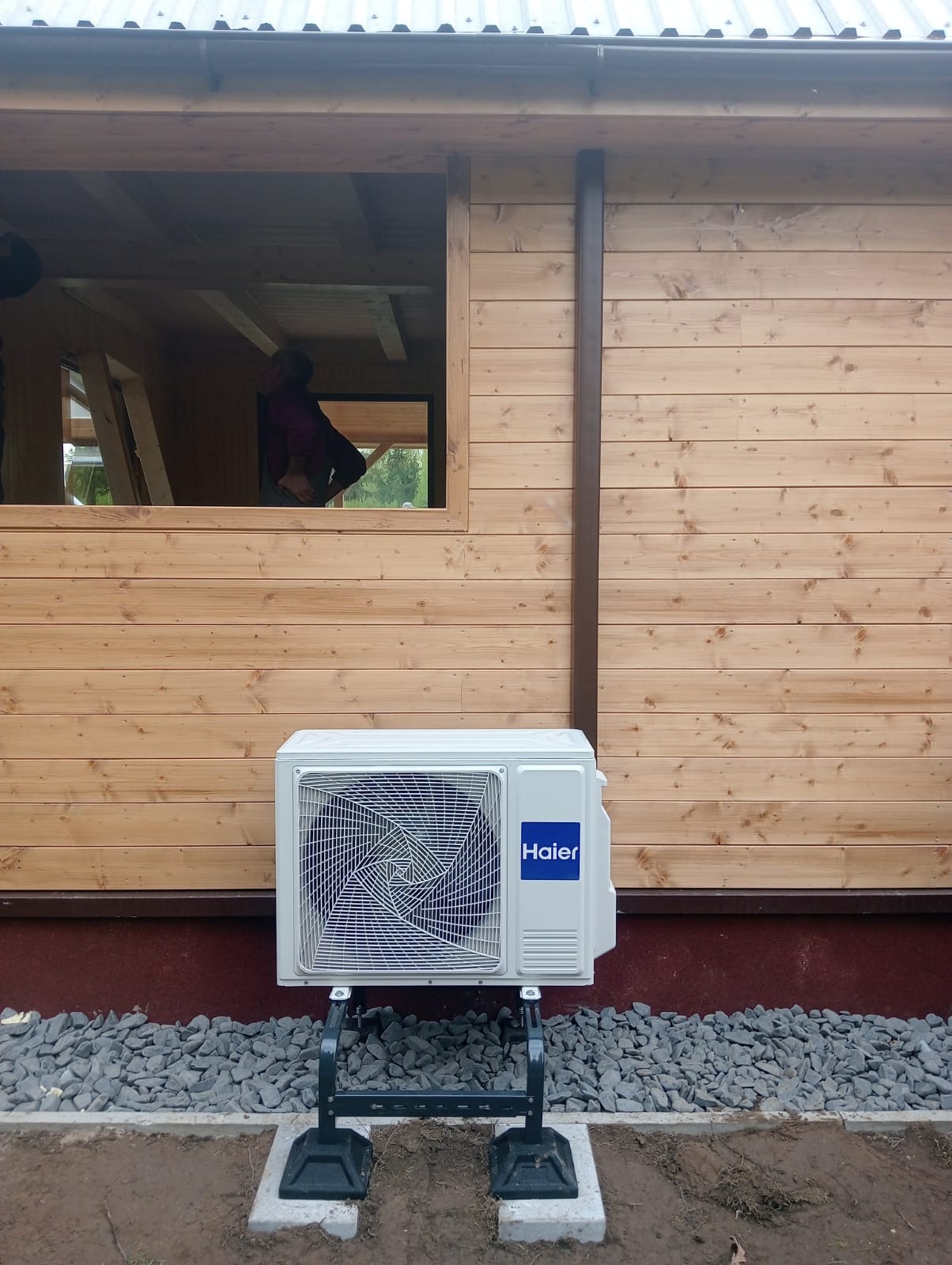 Biała jednostka zewnętrzna klimatyzacji Haier zamontowana na metalowym stelażu przed drewnianym domem w trakcie budowy, widoczna przez okno osoba wewnątrz.