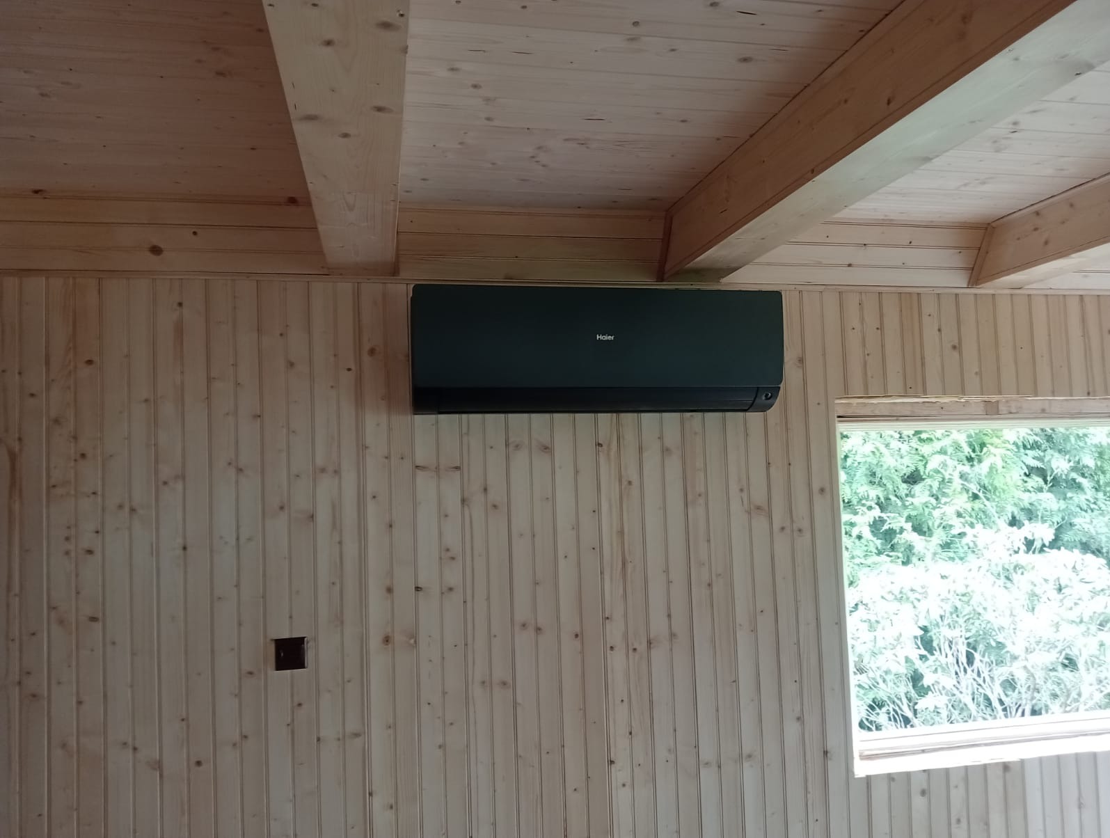 Czarny klimatyzator Haier zamontowany na drewnianej ścianie z boazerii w jasnym pomieszczeniu z drewnianym stropem i widokiem z okna na zieleń.