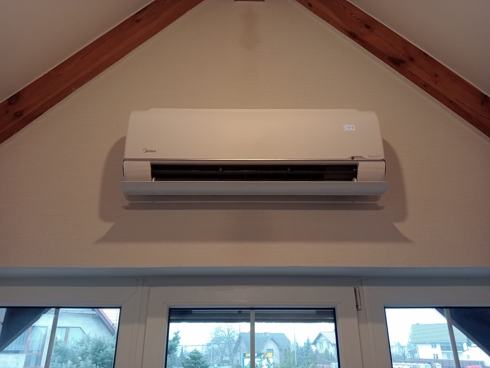 Biały klimatyzator Midea zamontowany w domu na poddaszu, widoczny na tle drewnianych belek stropowych i okna z widokiem na zabudowę jednorodzinną.