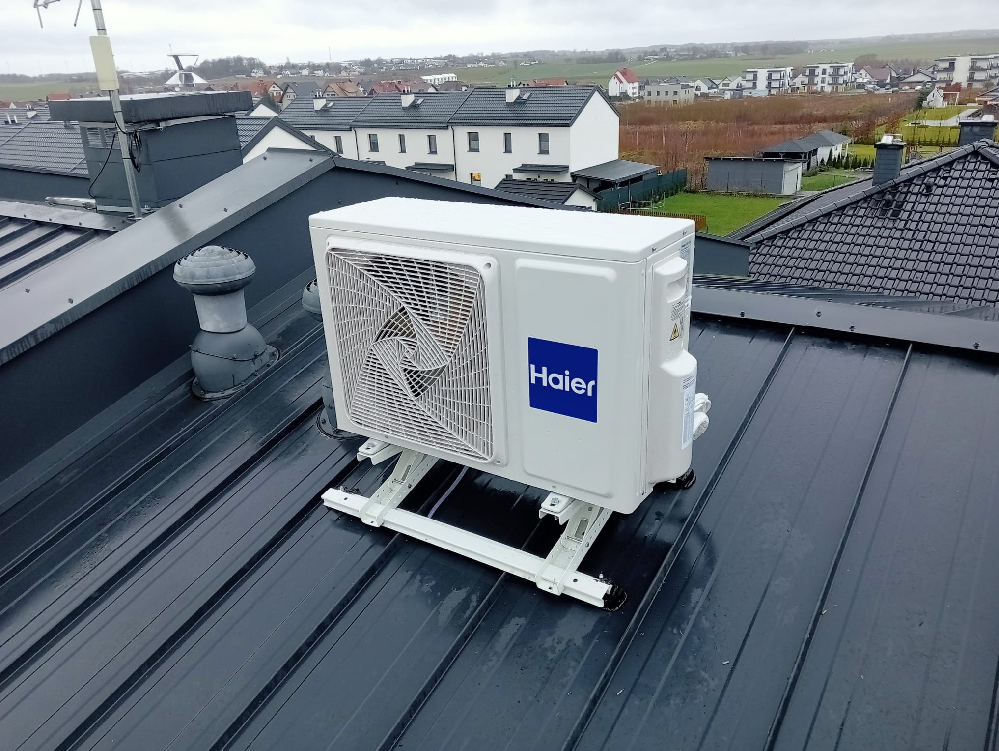 Zewnętrzna jednostka klimatyzacji Haier zamontowana na dachu domu jednorodzinnego, widok z góry na tle zabudowy mieszkalnej w pochmurny dzień.