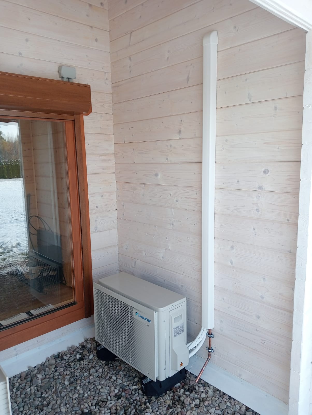 Zewnętrzna jednostka klimatyzacji Daikin zamontowana na elewacji domu z jasnego drewna, widoczne rury miedziane i biała maskownica.