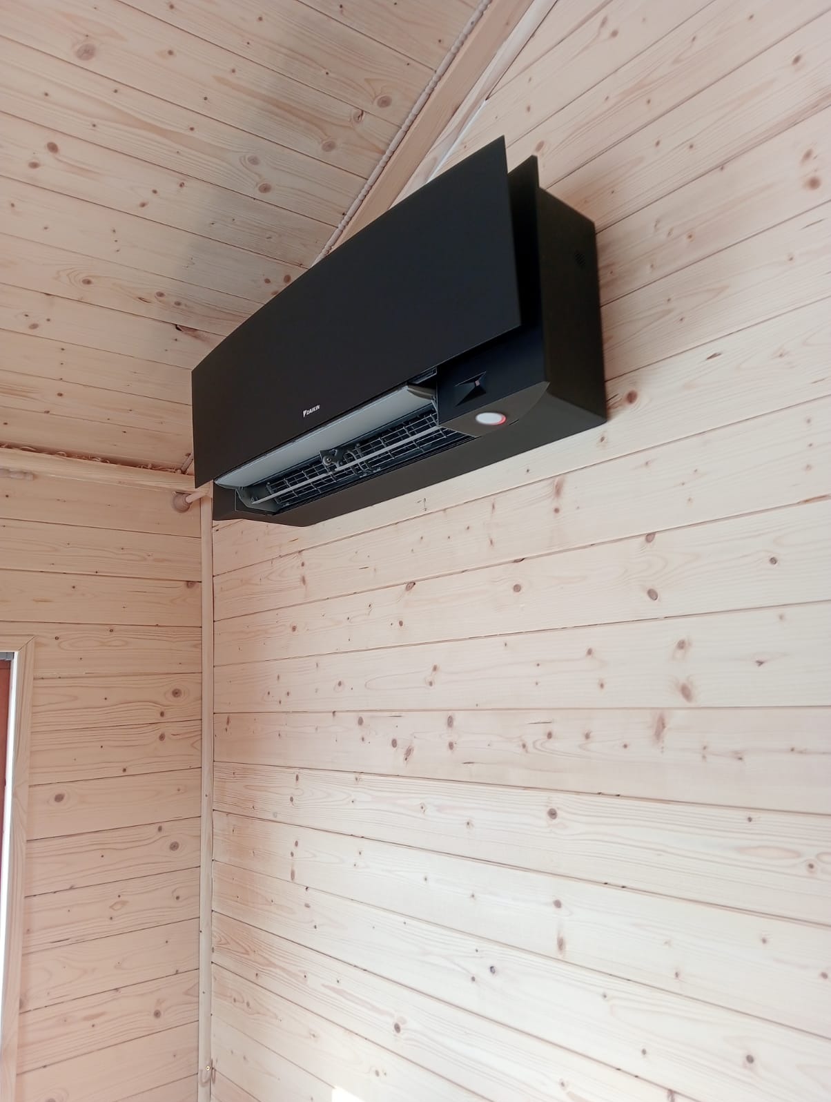 Czarna klimatyzacja Daikin zamontowana na ścianie z jasnego drewna w pomieszczeniu o wykończeniu drewnianym.