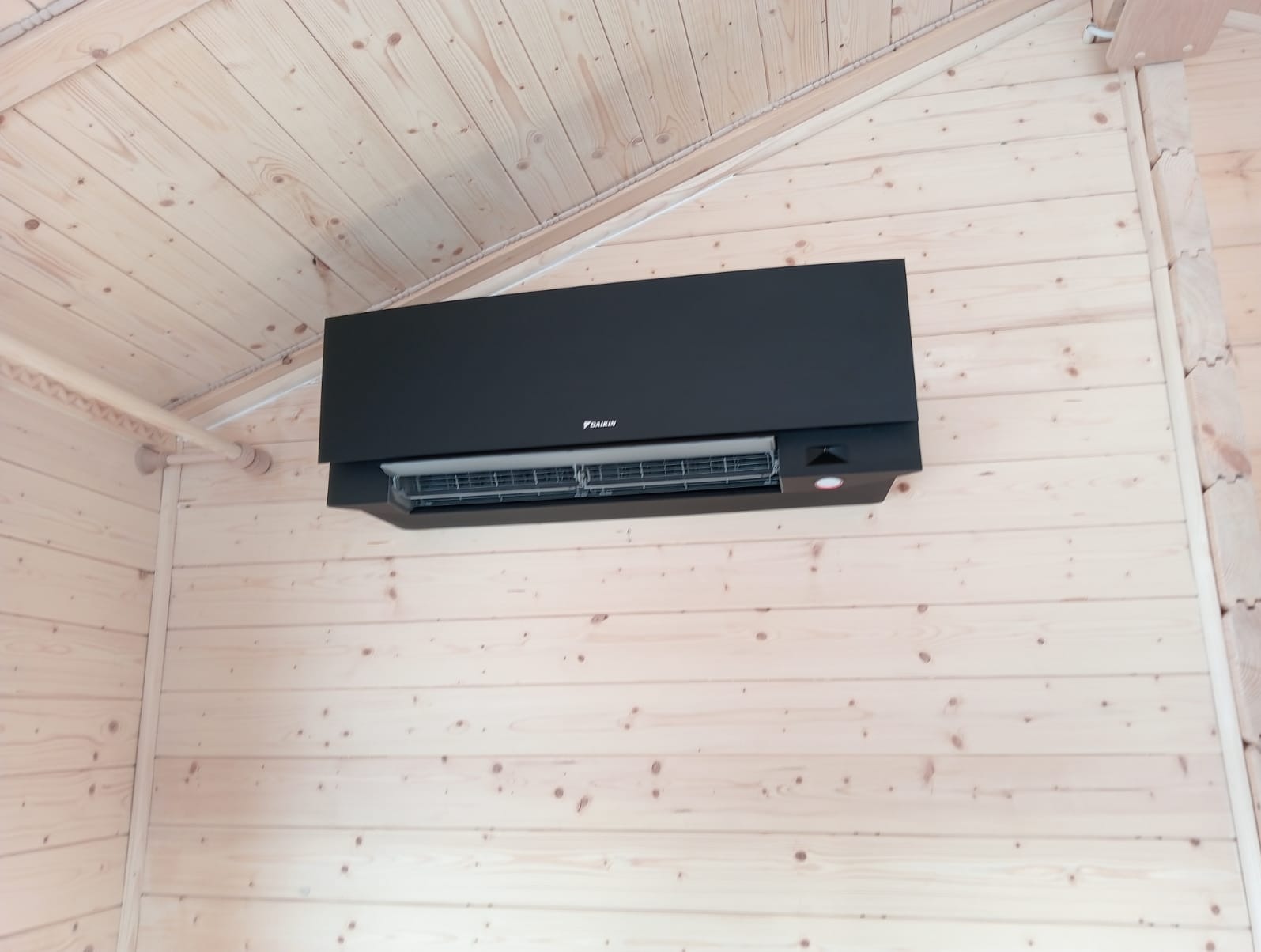 Czarna jednostka wewnętrzna klimatyzatora Daikin zamontowana na jasnej, drewnianej ścianie w pomieszczeniu o skośnym suficie wykończonym boazerią.