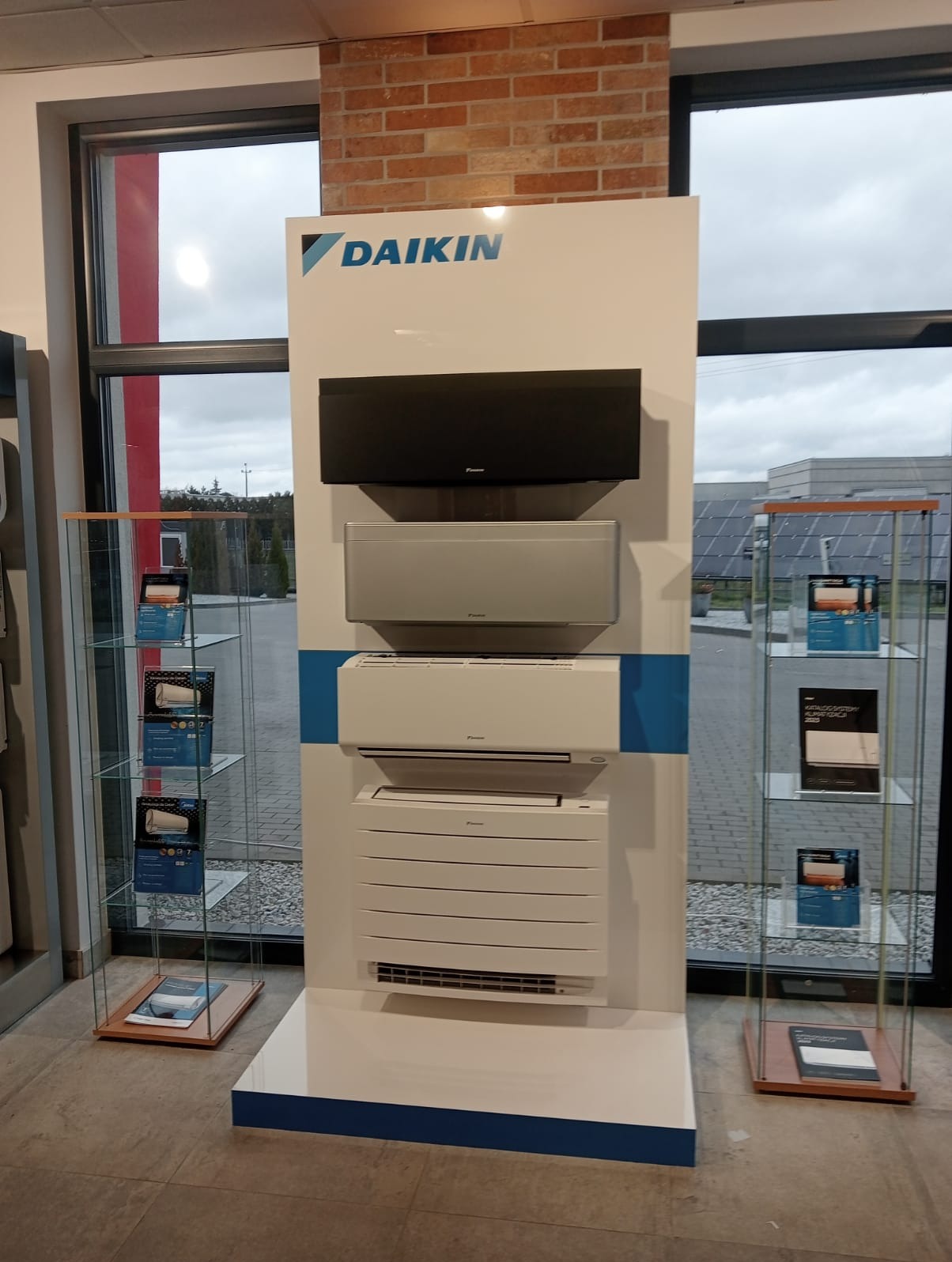 Ekspozycja klimatyzatorów Daikin w salonie sprzedaży, prezentująca modele ścienne w kolorach czarnym, srebrnym i białym, na tle ceglanej ściany i okna z widokiem na zewnątrz.