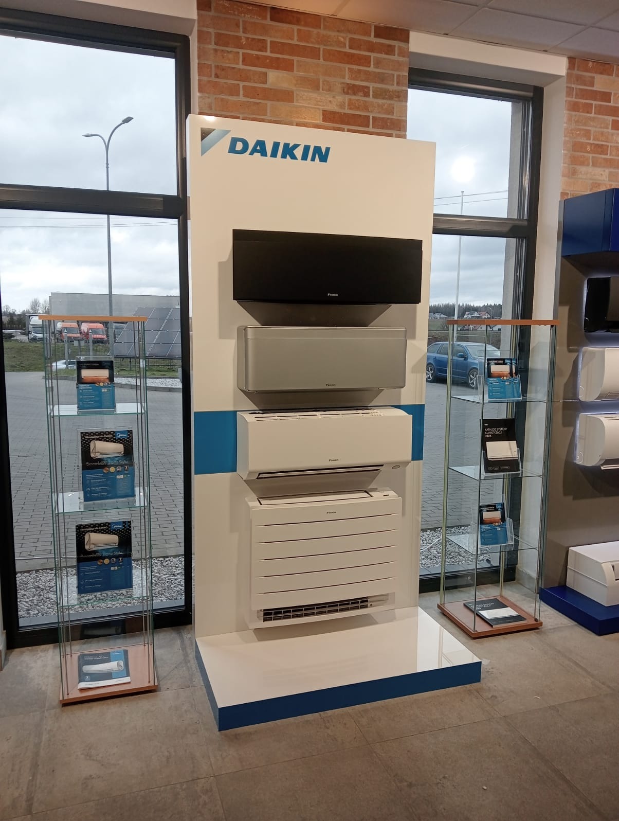 Ekspozycja klimatyzatorów Daikin w salonie: czarny, srebrny i biały model na białym panelu z niebieskim paskiem, obok przeszklone gabloty z katalogami, widok na parking za oknem.