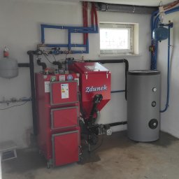 GRUPA MAJEWSCY SPÓŁKA Z OGRANICZONĄ ODPOWIEDZIALNOŚCIĄ - Czerwony piec na paliwo stałe marki Zdunek z zasobnikiem i szary zbiornik buforowy w kotłowni z niebieskimi rurami instalacji grzewczej.