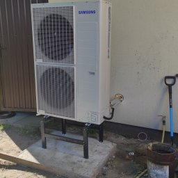 GRUPA MAJEWSCY SPÓŁKA Z OGRANICZONĄ ODPOWIEDZIALNOŚCIĄ - Zewnętrzna jednostka pompy ciepła Samsung, zamontowana na metalowym stelażu na betonowej podstawie przy ścianie budynku. Widoczne przewody i akcesoria instalacyjne, a także łopata oparta o ścianę.