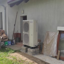 GRUPA MAJEWSCY SPÓŁKA Z OGRANICZONĄ ODPOWIEDZIALNOŚCIĄ - Zewnętrzna jednostka klimatyzacji Samsung, zamontowana na betonowych bloczkach przy ścianie budynku w Taczanowie Drugim. Widoczne rury przyłączeniowe i fragment trawnika.