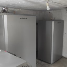 GRUPA MAJEWSCY SPÓŁKA Z OGRANICZONĄ ODPOWIEDZIALNOŚCIĄ - Dwie szare pompy ciepła Stalmark w piwnicy z betonową podłogą i sufitem, niebieska rura na suficie, surowa żarówka.