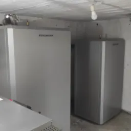 Dwie szare pompy ciepła Stalmark w piwnicy z betonową podłogą i sufitem, niebieska rura na suficie, surowa żarówka.