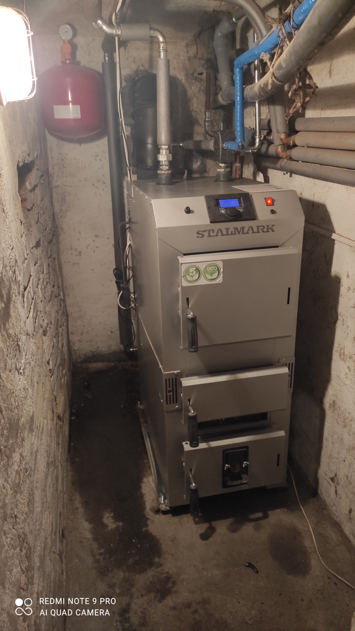 Kocioł grzewczy marki Stalmark w piwnicy z widocznymi rurami instalacji, zbiornikiem wyrównawczym i wilgotną podłogą.