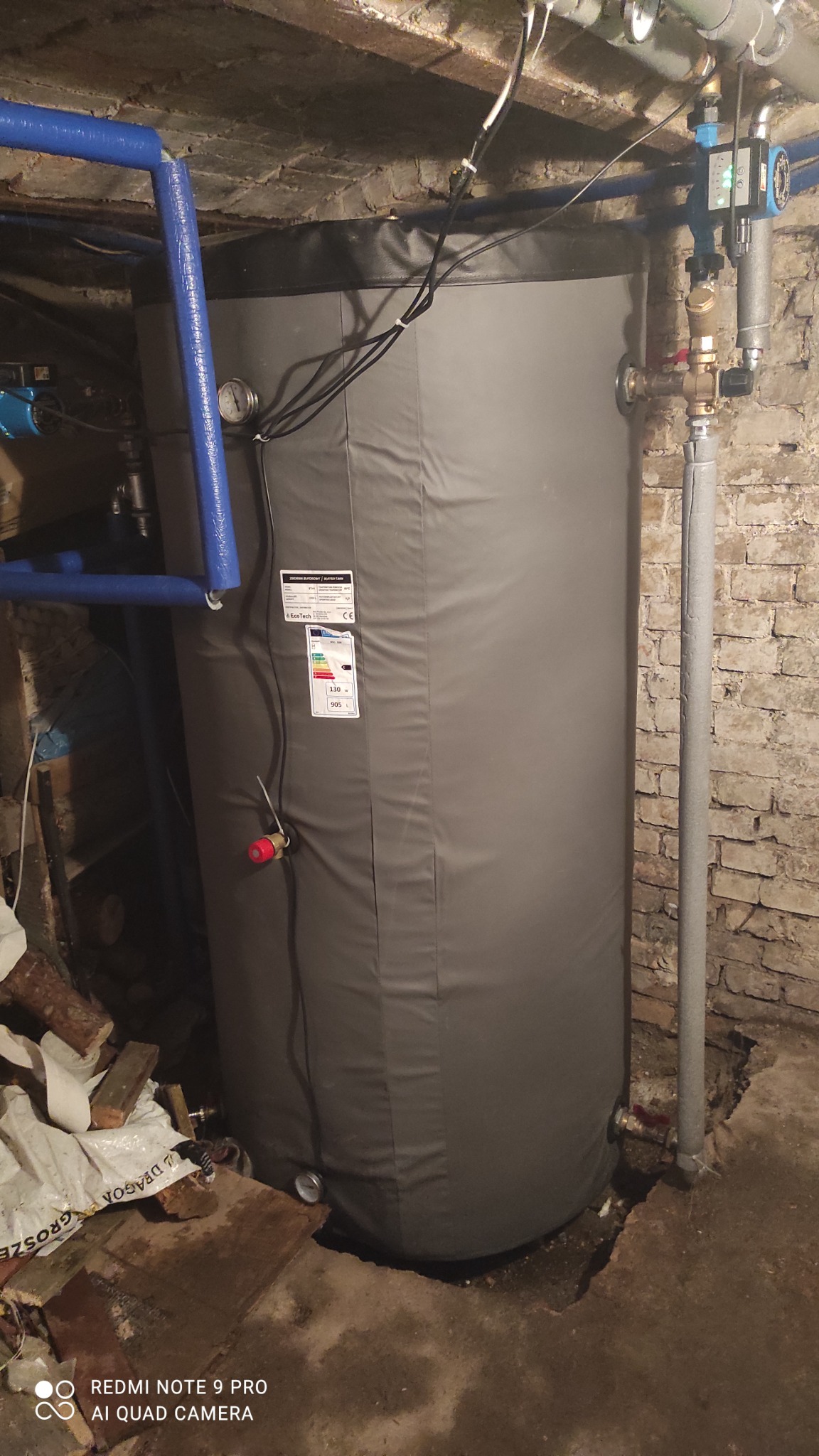 Szary, cylindryczny zbiornik buforowy z etykietą energetyczną w piwnicy z odsłoniętą cegłą, widoczne rury w izolacji i fragmenty drewna na podłodze.