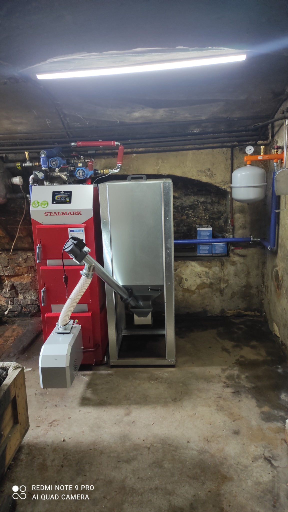 Czerwony piec na paliwo stałe marki Stalmark z podajnikiem, zainstalowany w piwnicy, widoczne rury i zbiornik wyrównawczy.