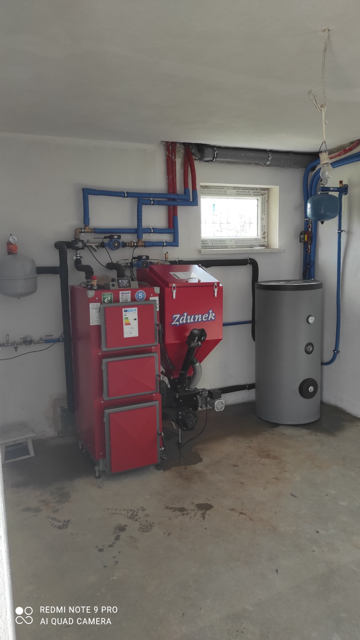 Czerwony piec na paliwo stałe marki Zdunek z zasobnikiem i szary zbiornik buforowy w kotłowni z niebieskimi rurami instalacji grzewczej.