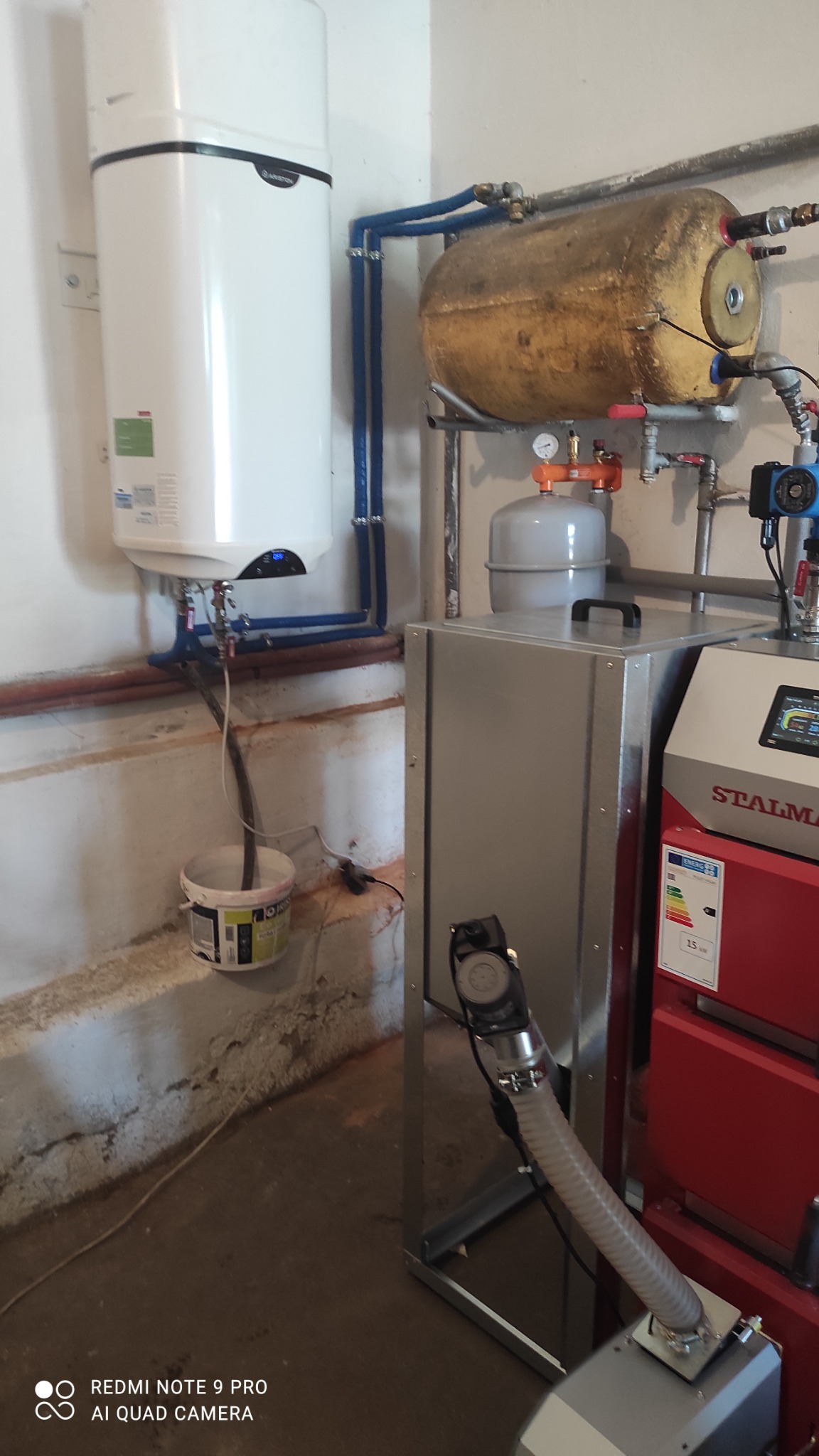 Biały zasobnik ciepłej wody użytkowej Ariston, brązowy zbiornik buforowy, szary zbiornik wyrównawczy, oraz czerwony piec Stalmark z podajnikiem ślimakowym w kotłowni