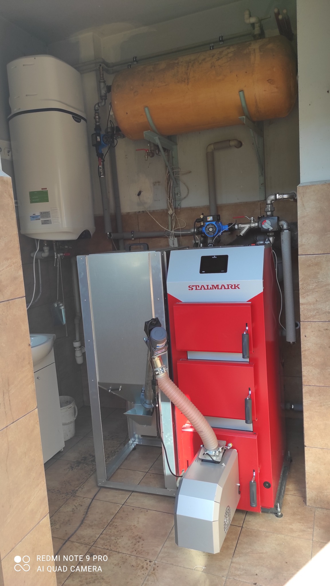 Czerwony piec Stalmark z podajnikiem, zbiornikiem na wodę i zasobnikiem buforowym w kotłowni, widoczne elementy instalacji hydraulicznej.