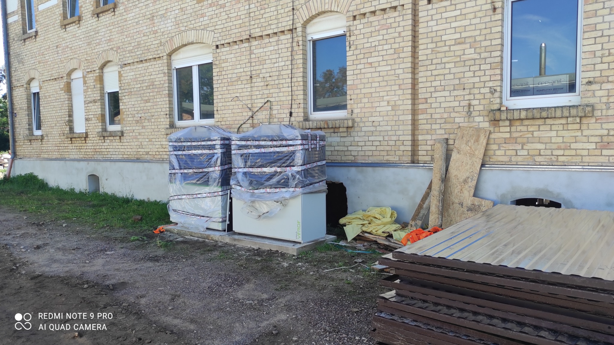 Dwie jednostki zewnętrzne, prawdopodobnie systemów grzewczych, zabezpieczone folią, stoją na zewnątrz budynku z cegły, obok sterty blachy falistej i innych materiałów budowlanych.