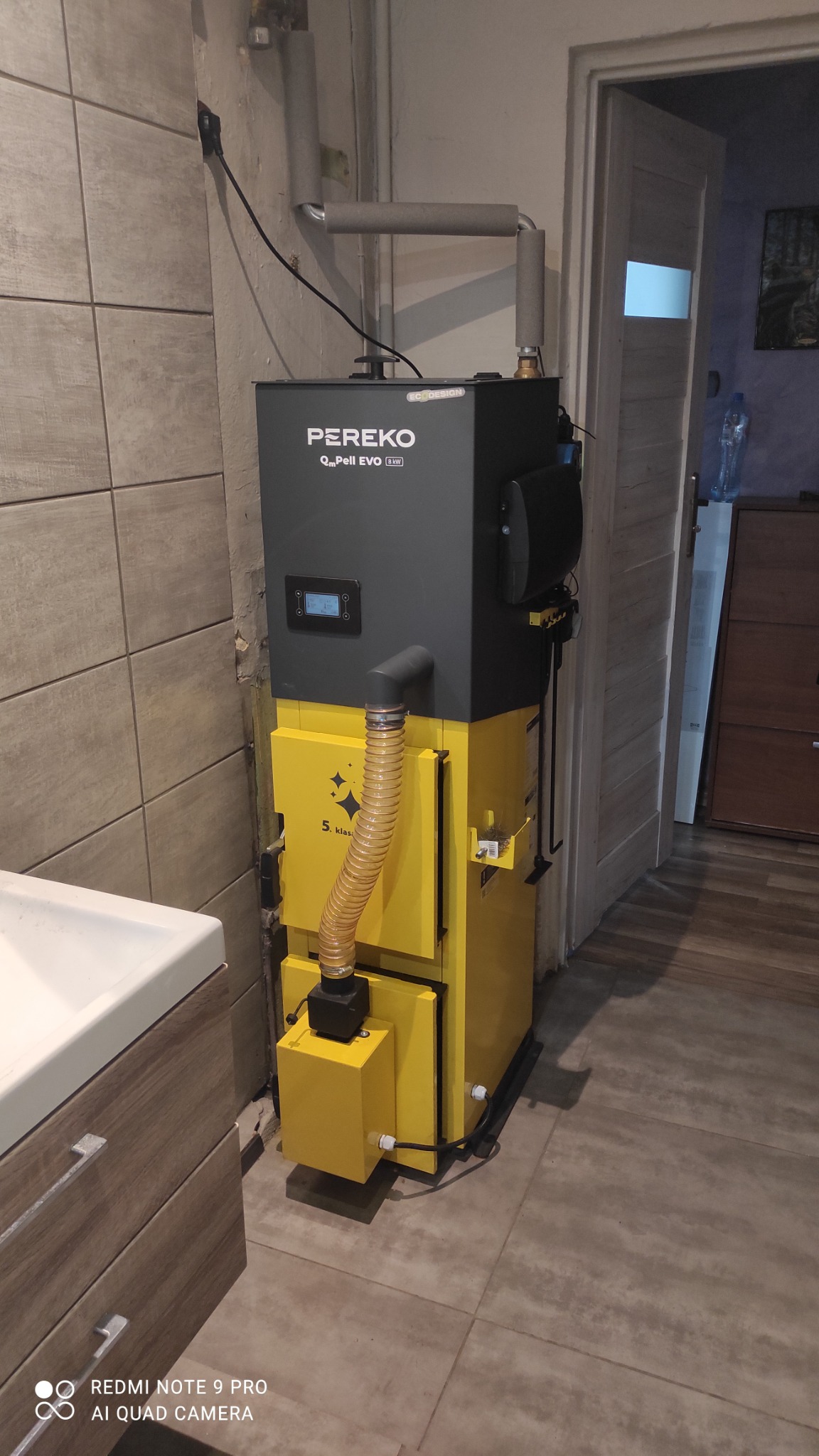 Żółto-szary piec na pellet marki PEREKO QmPell EVO 8 kW, z podajnikiem i rurą spiro, zainstalowany w kotłowni z beżowymi płytkami i białą umywalką z szafką.