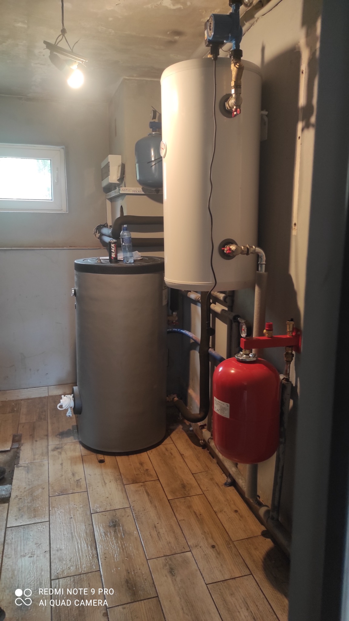 Dwa zbiorniki na wodę: szary i biały, połączone rurami, z czerwonym zbiornikiem wyrównawczym. Na szarym zbiorniku stoją napoje. Widok na pomieszczenie z drewnopodobną podłogą i oknem.