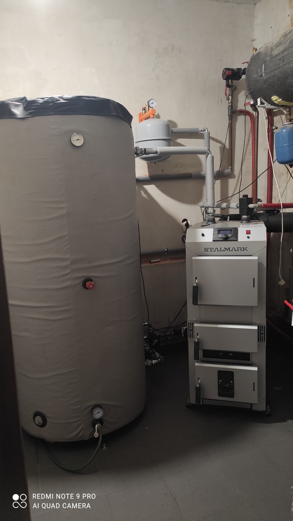 Stalmark piec z podajnikiem, stojący obok dużego zbiornika akumulacyjnego w kotłowni o surowych ścianach.