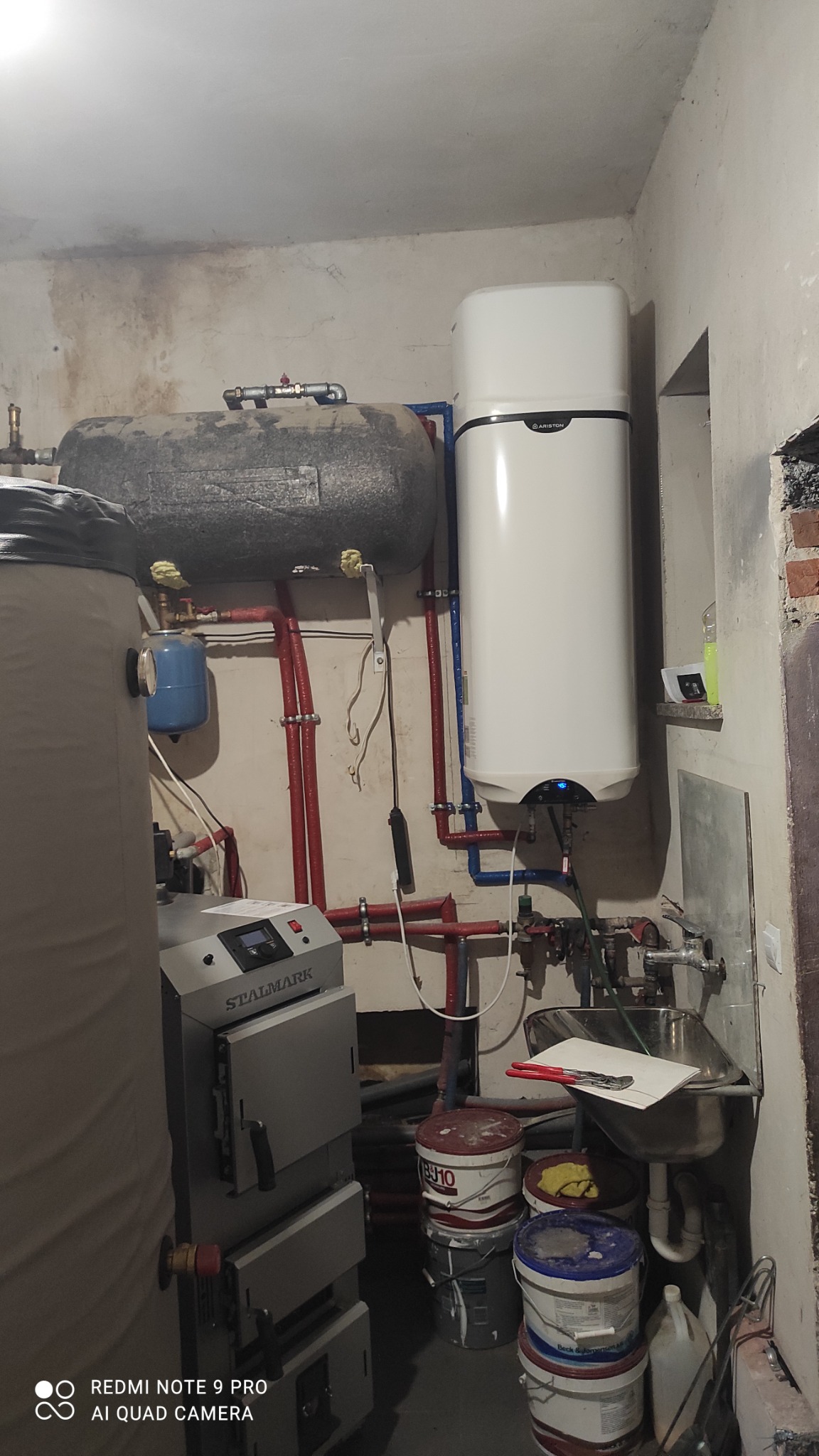 Kotłownia z piecem STALMARK, zbiornikiem akumulacyjnym, bojlerem Ariston i instalacją hydrauliczną z czerwonymi i niebieskimi rurami, widoczne wiaderka farb i umywalka