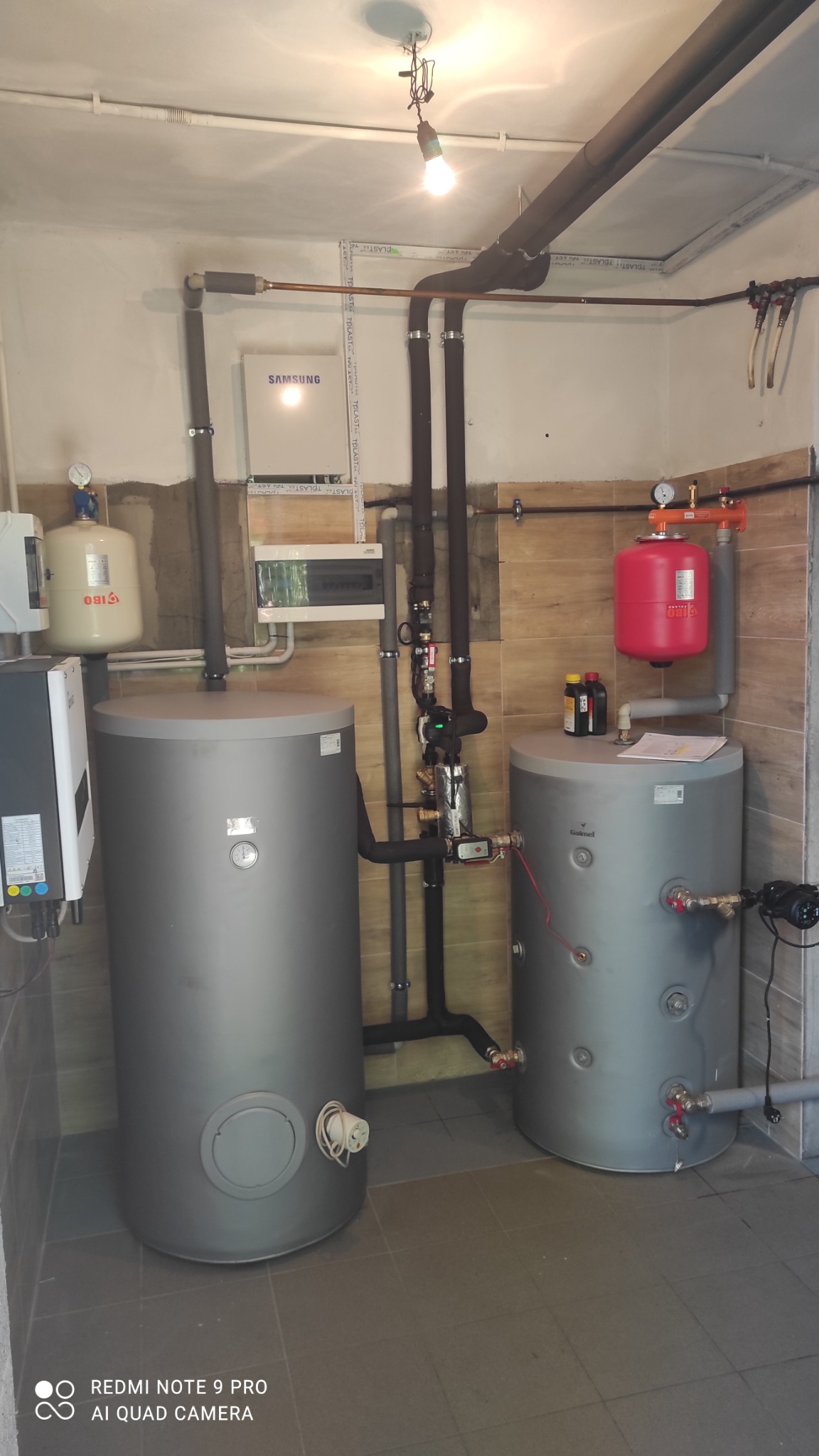 Dwa szare zbiorniki na wodę z podłączonymi rurami i osprzętem w kotłowni, z widocznym bojlerem IBO, panelem Samsung i czerwonym zbiornikiem Galmet. Widoczne także butelki z płynem niezamarzającym.