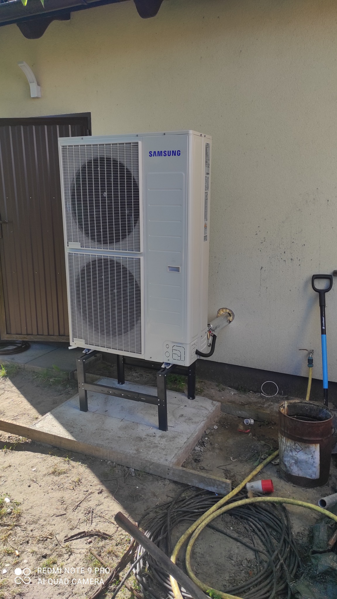 Zewnętrzna jednostka pompy ciepła Samsung, zamontowana na metalowym stelażu na betonowej podstawie przy ścianie budynku. Widoczne przewody i akcesoria instalacyjne, a także łopata oparta o ścianę.