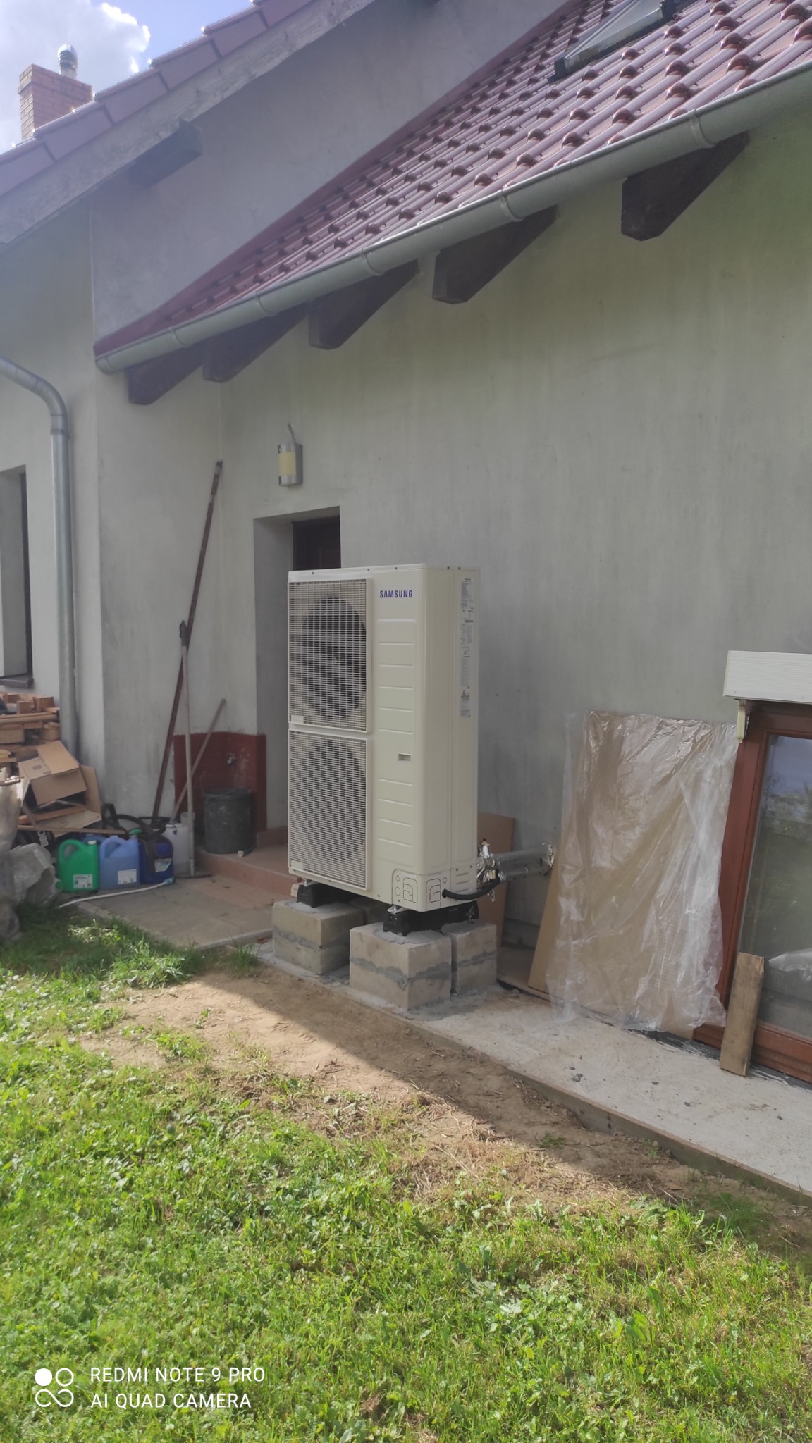 Zewnętrzna jednostka klimatyzacji Samsung, zamontowana na betonowych bloczkach przy ścianie budynku w Taczanowie Drugim. Widoczne rury przyłączeniowe i fragment trawnika.